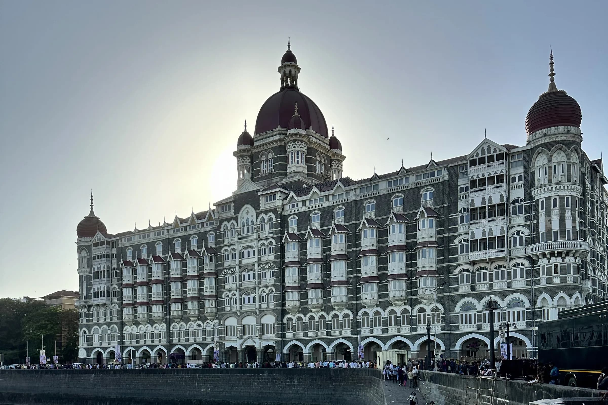 El ala del Palacio de The Taj Mahal Palace con sus cúpulas rojas, vista al otro lado del puerto, con pequeñas barcas en el muelle