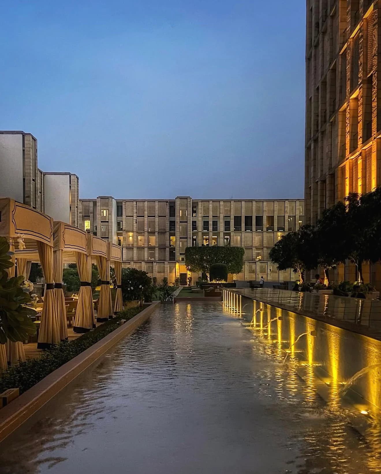 The Lodhi, en Nueva Delhi, al anochecer: la larga piscina con fuentes iluminadas, cabañas con cortinas y las alas de arenisca del hotel