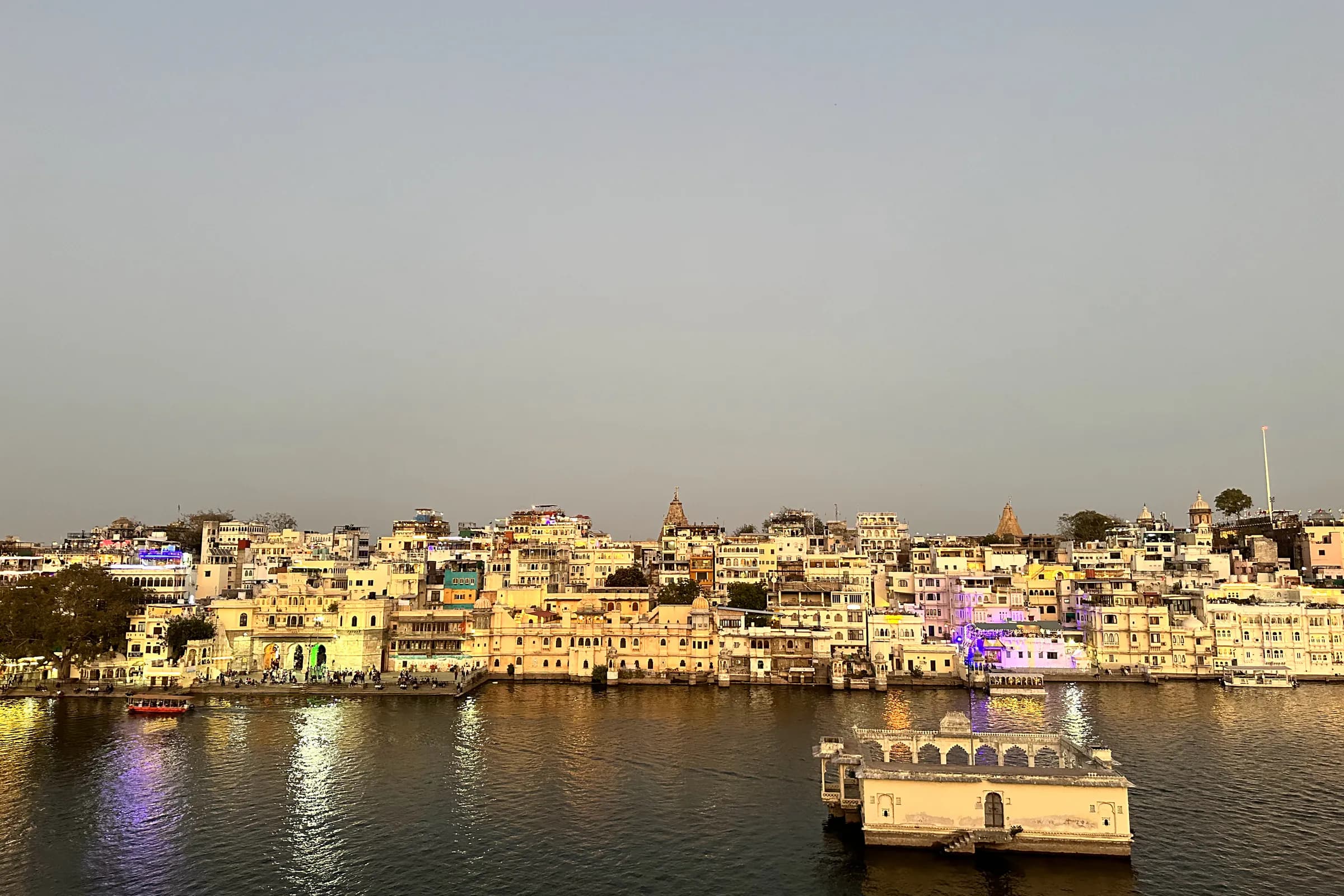 El casco antiguo de Udaipur a orillas del lago Pichola al anochecer, con las luces encendiéndose en la orilla y una barca iluminada sobre el agua