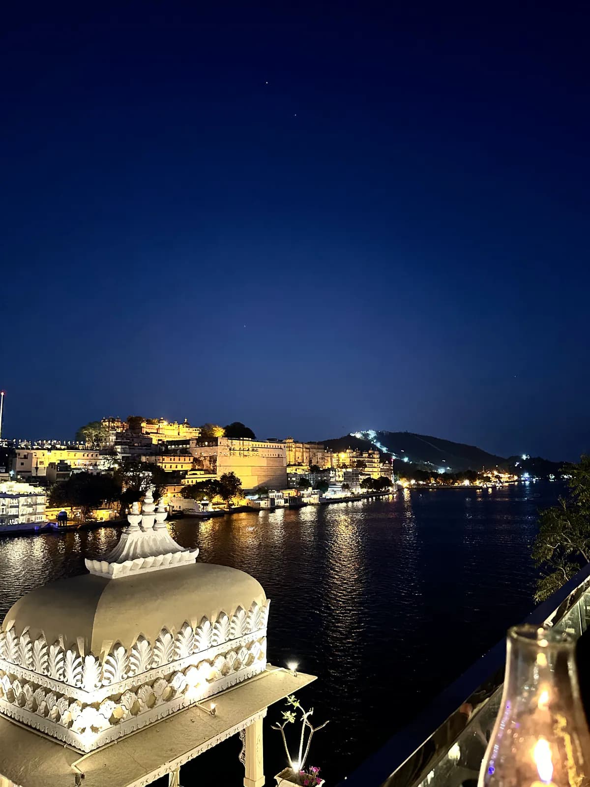 El lago Pichola de noche desde la azotea de Upre by 1559 AD, con una cúpula blanca tallada en primer plano y el Palacio de la Ciudad iluminado al otro lado del agua