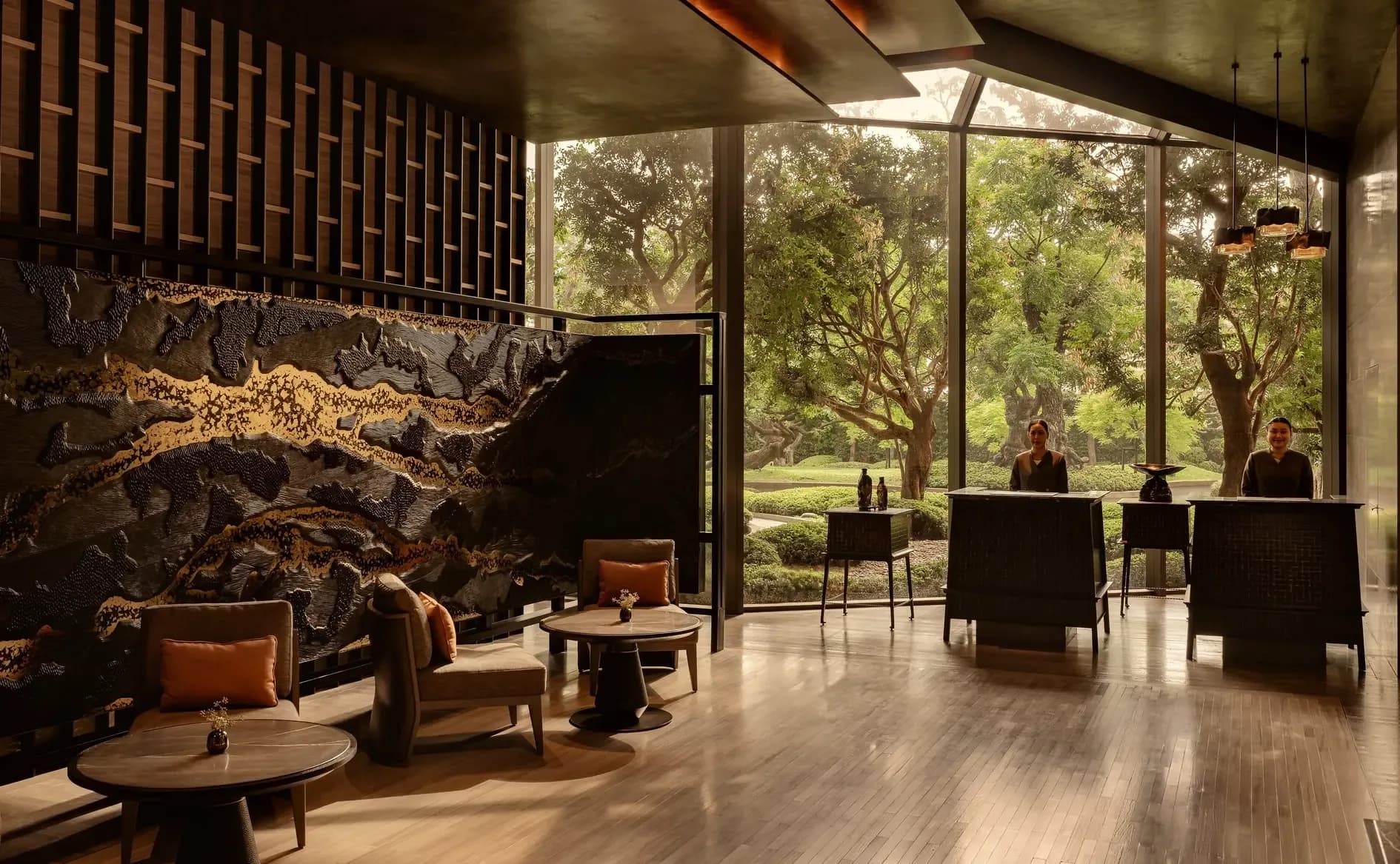 The residence lobby at Aman Nai Lert Bangkok, lounge seating beneath a timber ceiling, a dark mineral feature wall, and full-height glass onto the mature trees of the Nai Lert park.