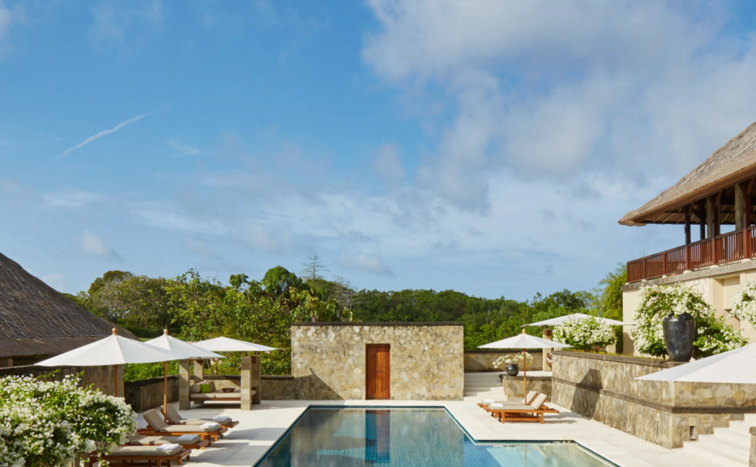 A private pool terrace at Aman Villas at Nusa Dua, parasols and daybeds on the stone deck, a thatched bale to one side and the ravine forest beyond.