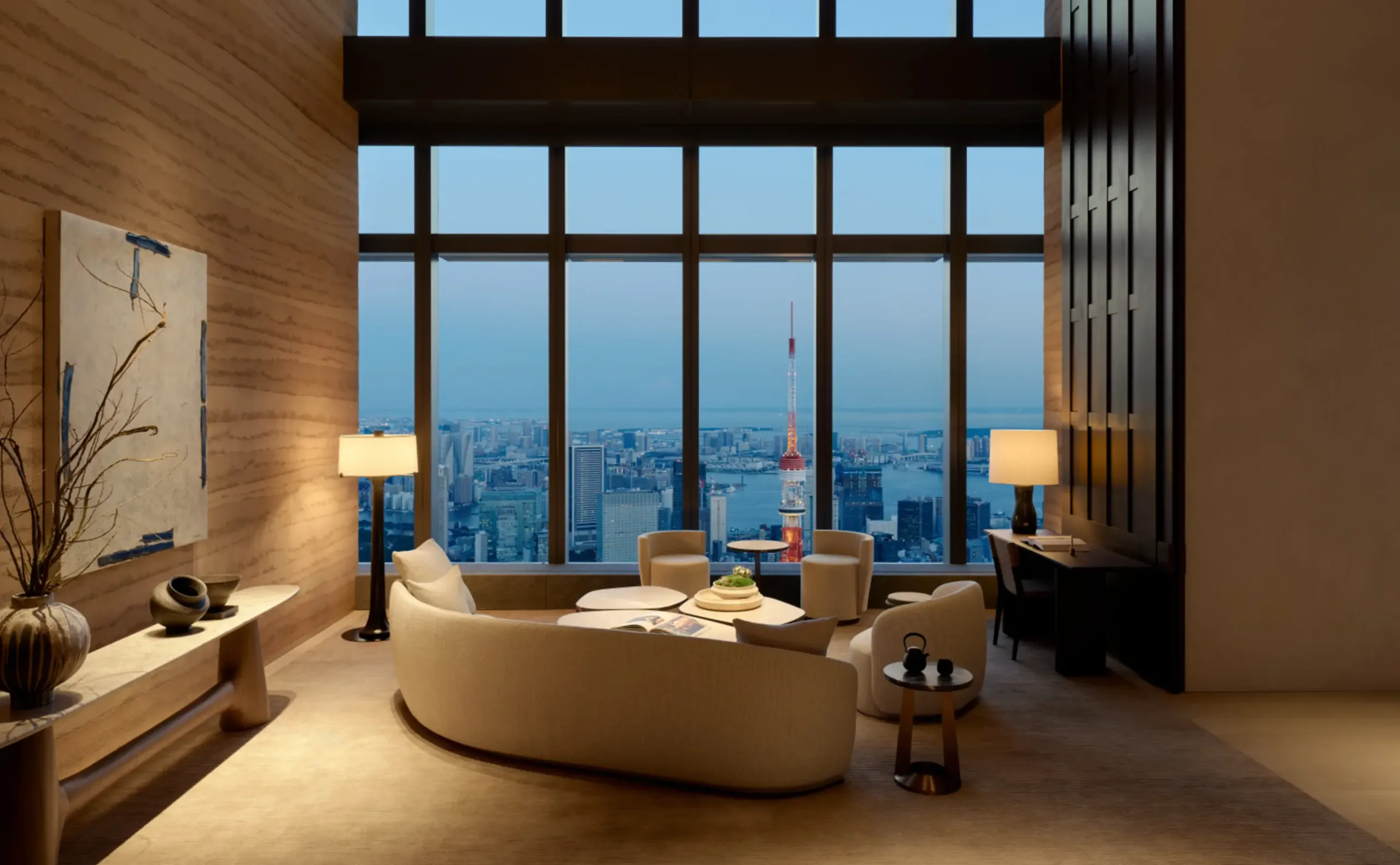 The owners' library at Aman Residences, Tokyo, a curved sofa and armchairs against stone walls, with Tokyo Tower lit beyond the full-height windows at dusk.