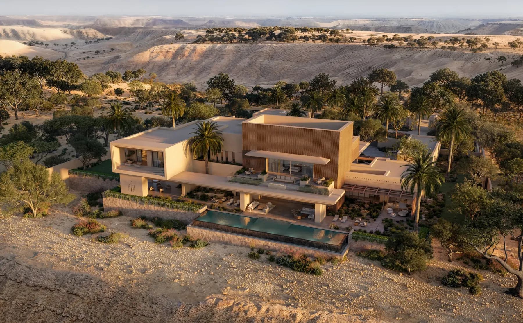 An aerial view at dusk of a single flat-roofed stone residence stepping down a wadi escarpment at Amansamar, a long pool on its lower terrace and date palms around it.