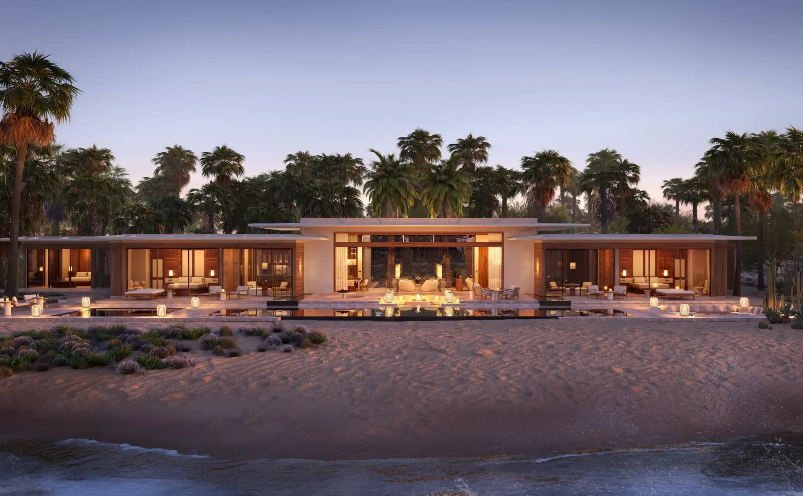 A beachfront villa at Aman Residences, Amanvari. A single-storey pavilion lit from within at dusk, set back from the sand with palms rising behind it.