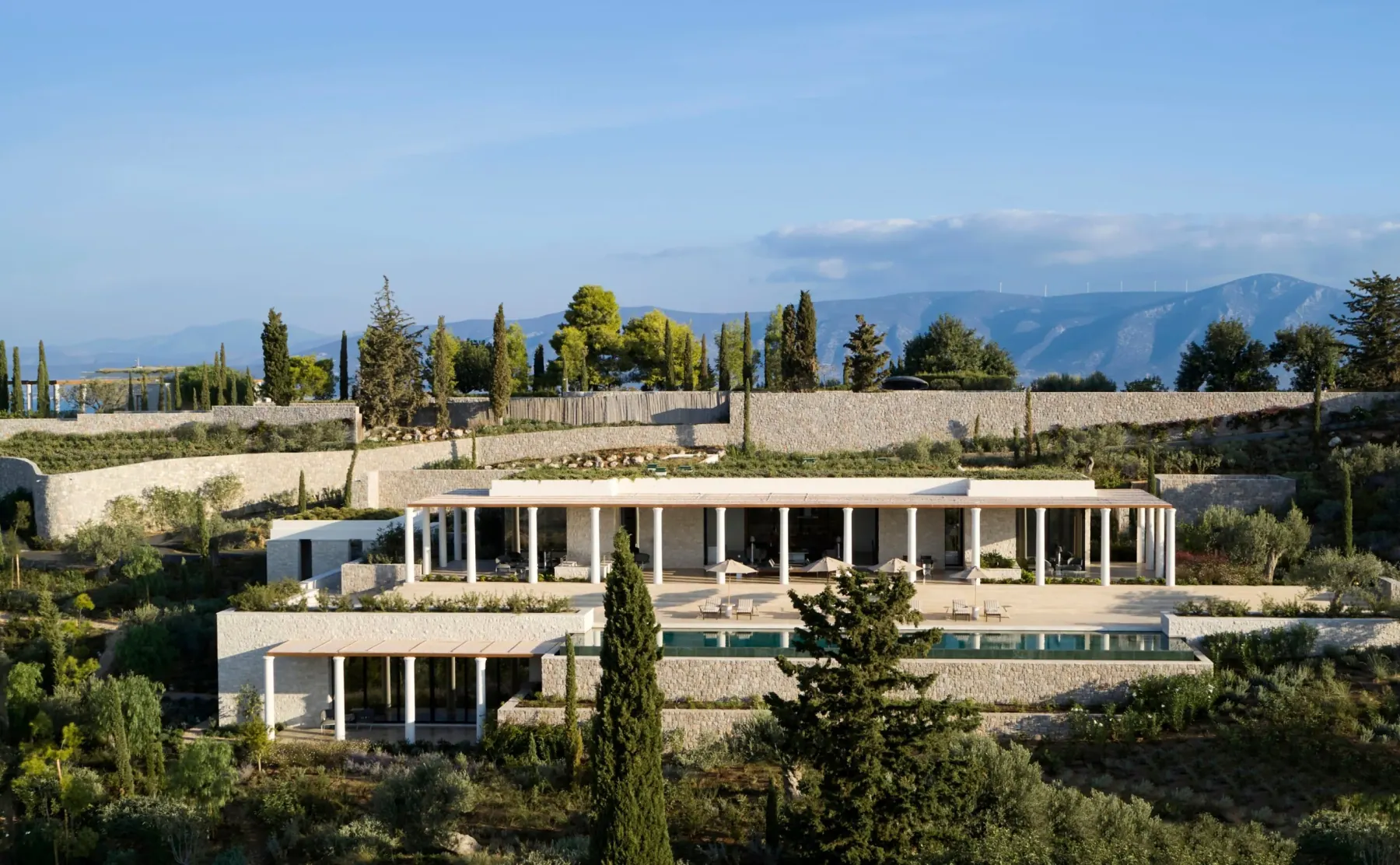 Villa 22 at Amanzoe seen from the neighbouring hillside, a white colonnaded villa terracing down to its own pool, with cypresses and the Argolic hills behind it.
