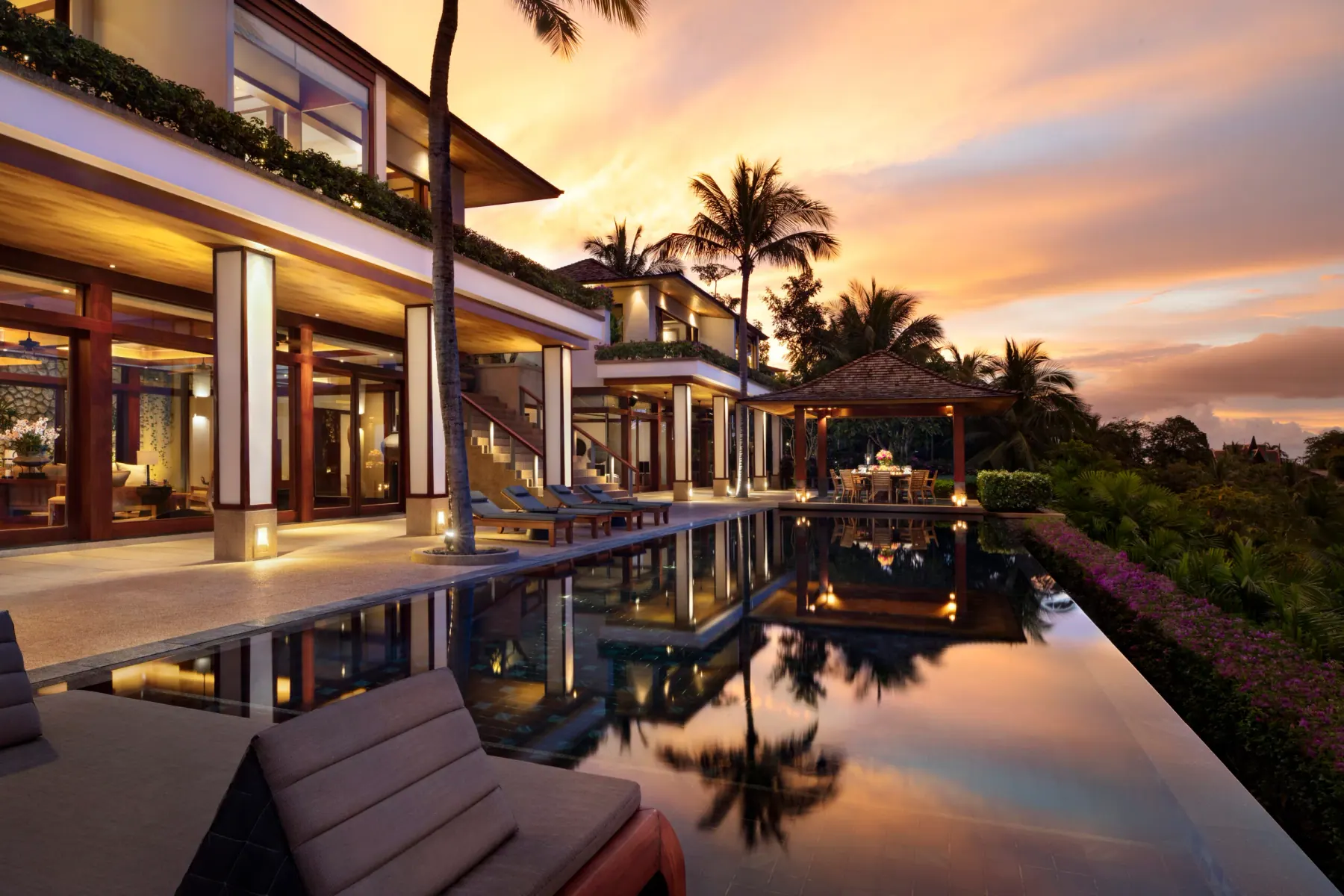 A pool villa at Andara at dusk, its terrace lit beneath deep eaves and reflected the length of a private pool, with palms and the Andaman Sea beyond.