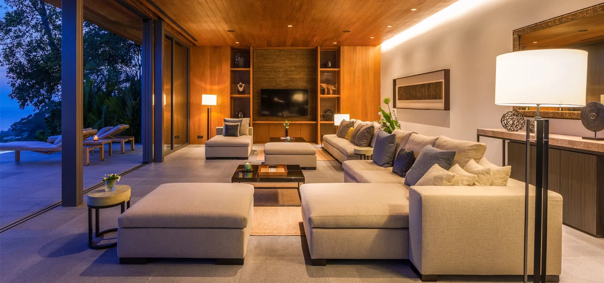 Open-plan villa living room at dusk, sectional sofa, sliding glass wall open to sea-view pool terrace