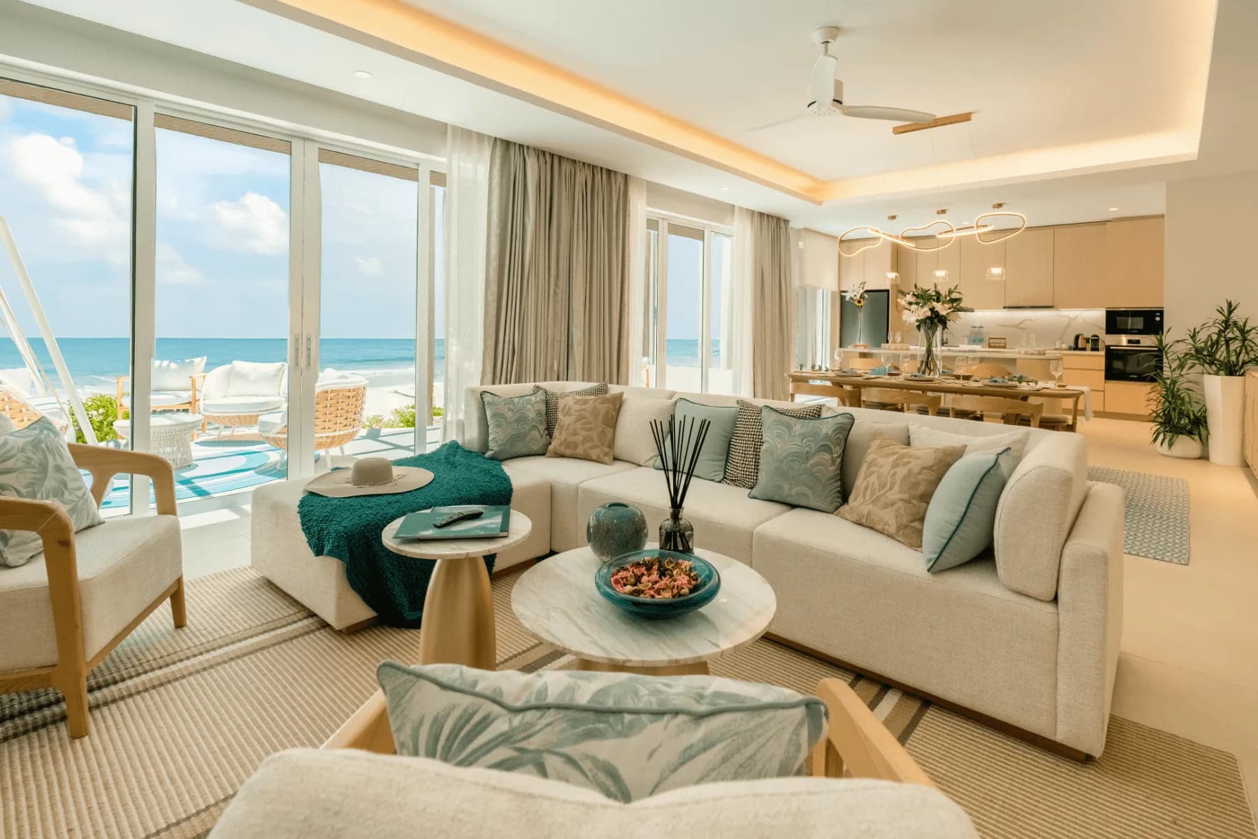 The living room of a Coral Residence at Kandima, a curved sofa and armchairs facing full-height glass, with the open kitchen behind and the lagoon past the terrace.