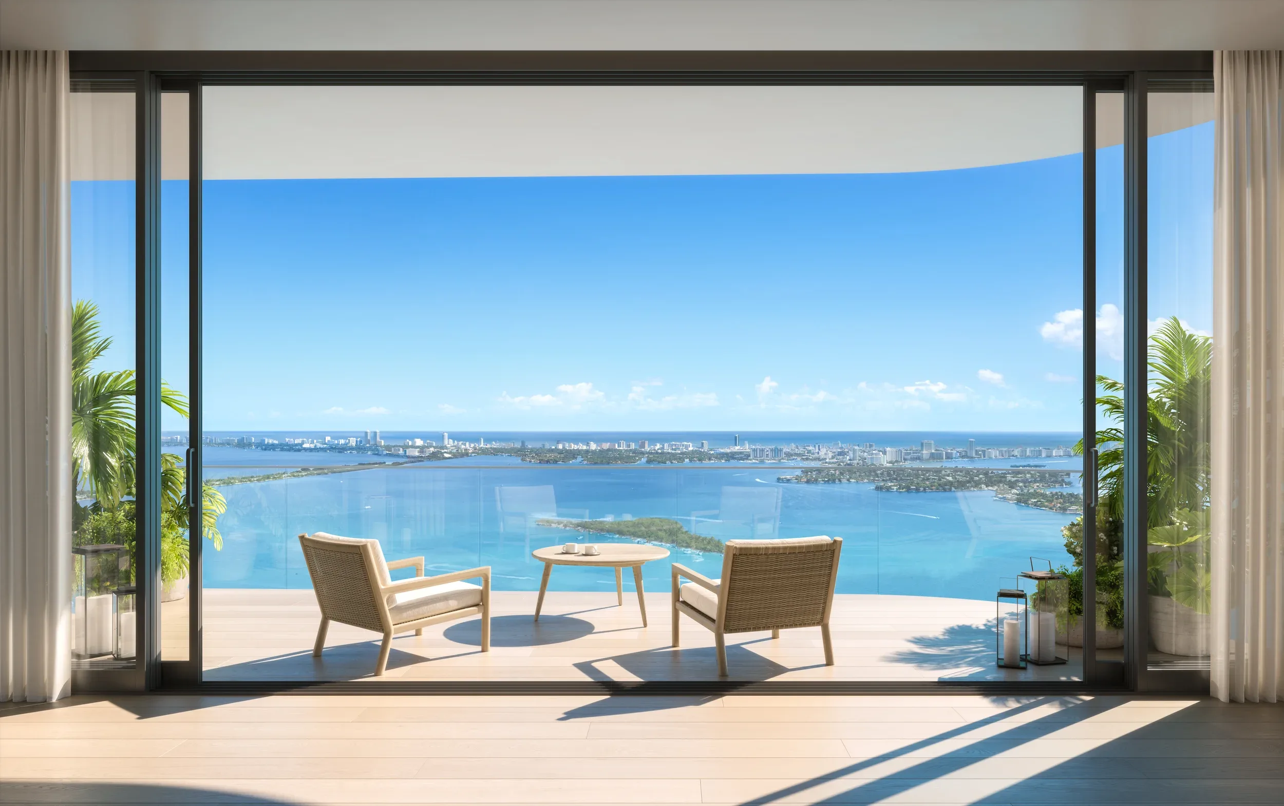 The terrace of a residence at EDITION Residences Miami Edgewater, its glass wall drawn back to a shaded deck with armchairs and a low table, looking west across Biscayne Bay.