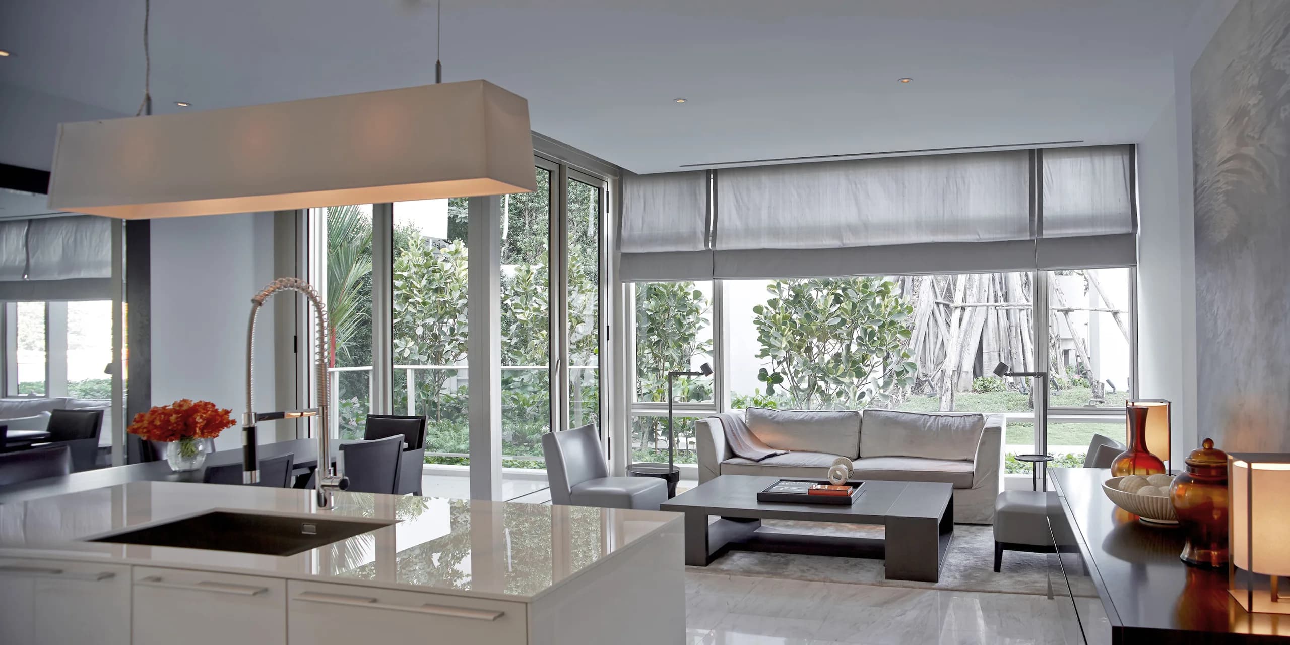 A residence interior at Four Seasons Private Residences Bangkok. An open kitchen island giving onto the living and dining room, with Roman blinds drawn back from a planted outlook.