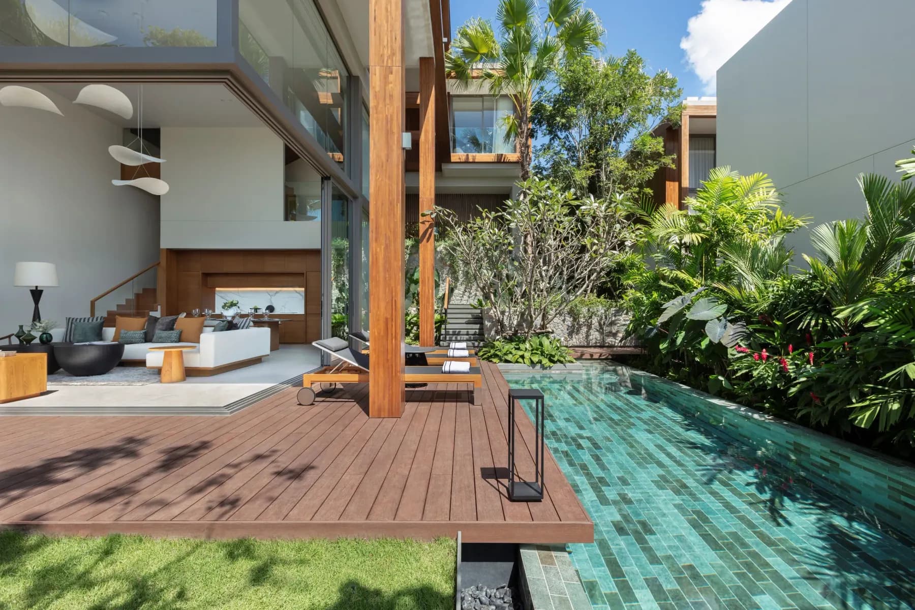 Pool terrace of a three-bedroom villa: timber deck and loungers beside a green-tiled pool and dense planting, the double-height living room open behind