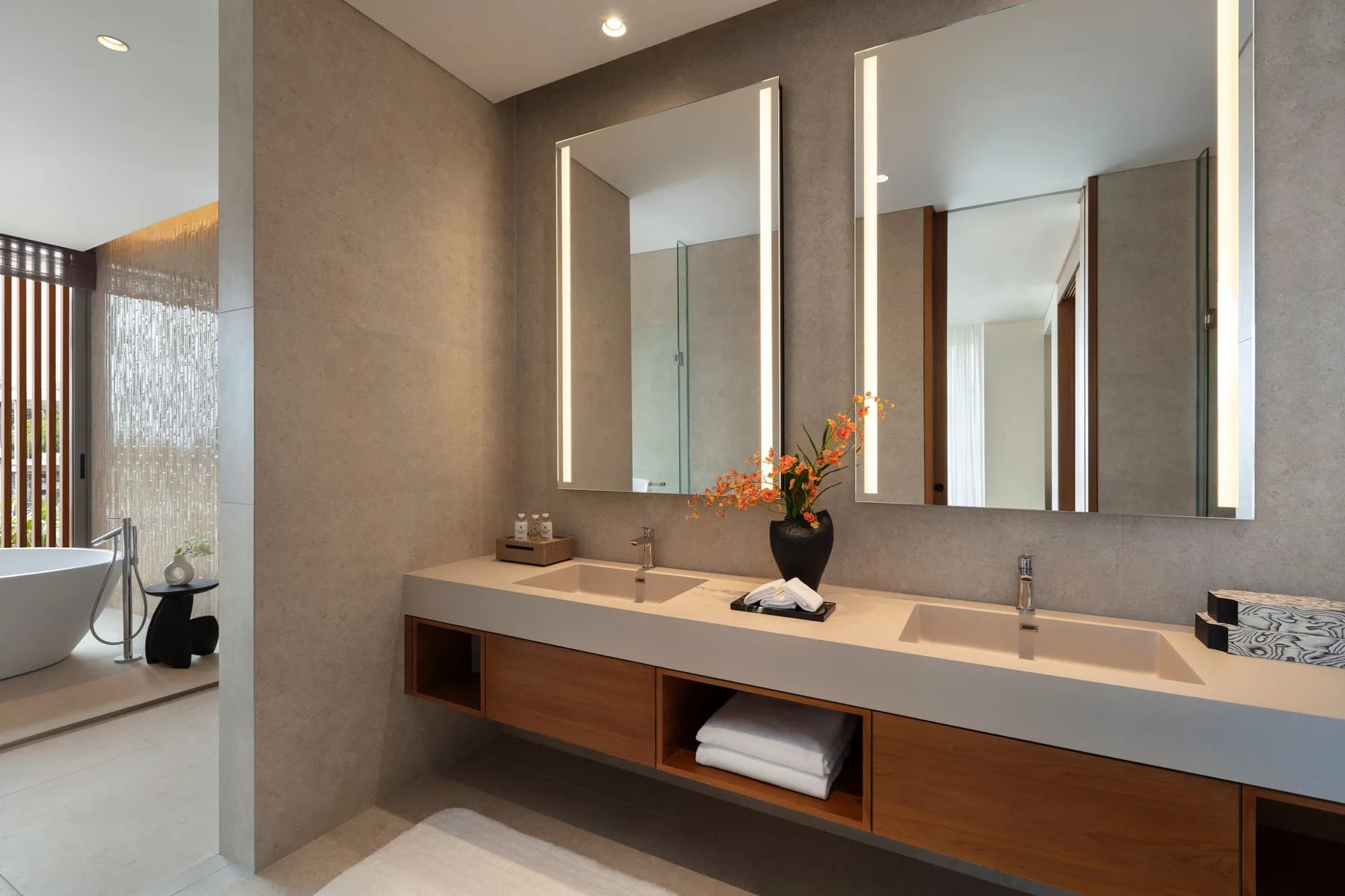 Bathroom with a double vanity under two lit mirrors and a freestanding bath beside a screened window