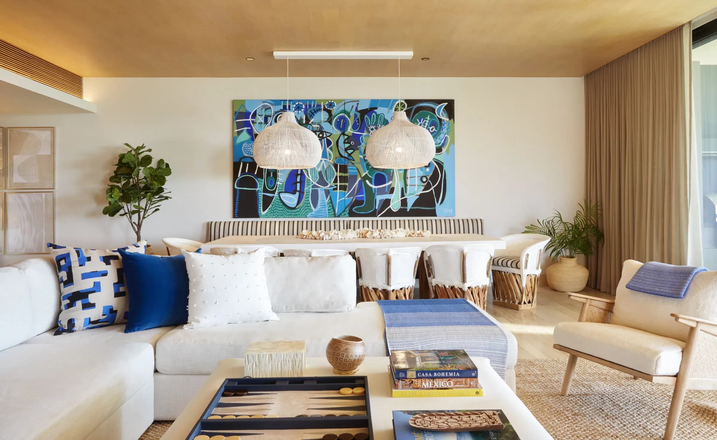 The living room of a residence at Montage Los Cabos, a long pale sofa beneath a large abstract canvas, woven pendant lights and a low table stacked with books.