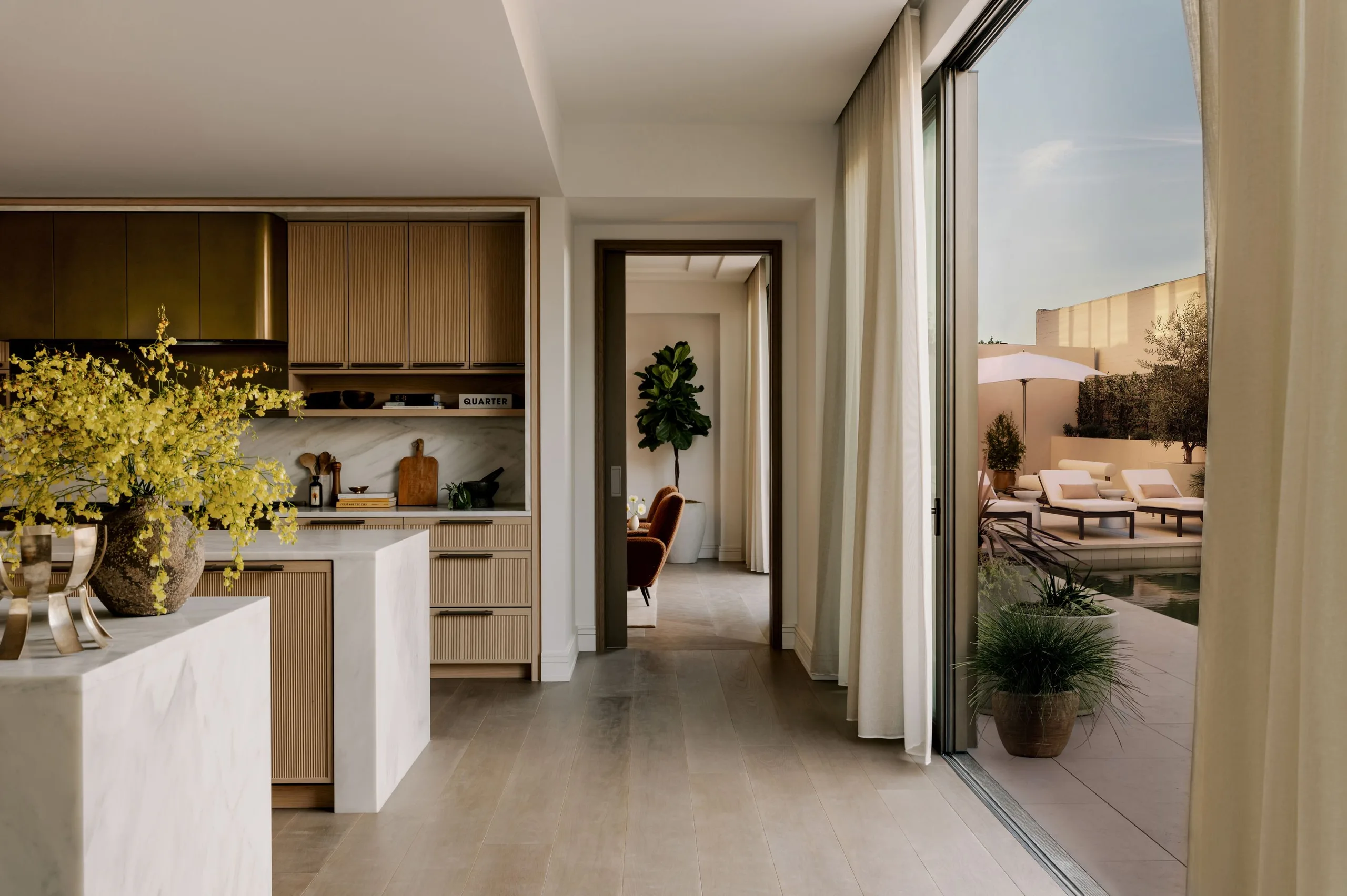 A residence interior at Rosewood Residences Beverly Hills. A timber kitchen and marble island, with sliding doors open to a planted terrace.