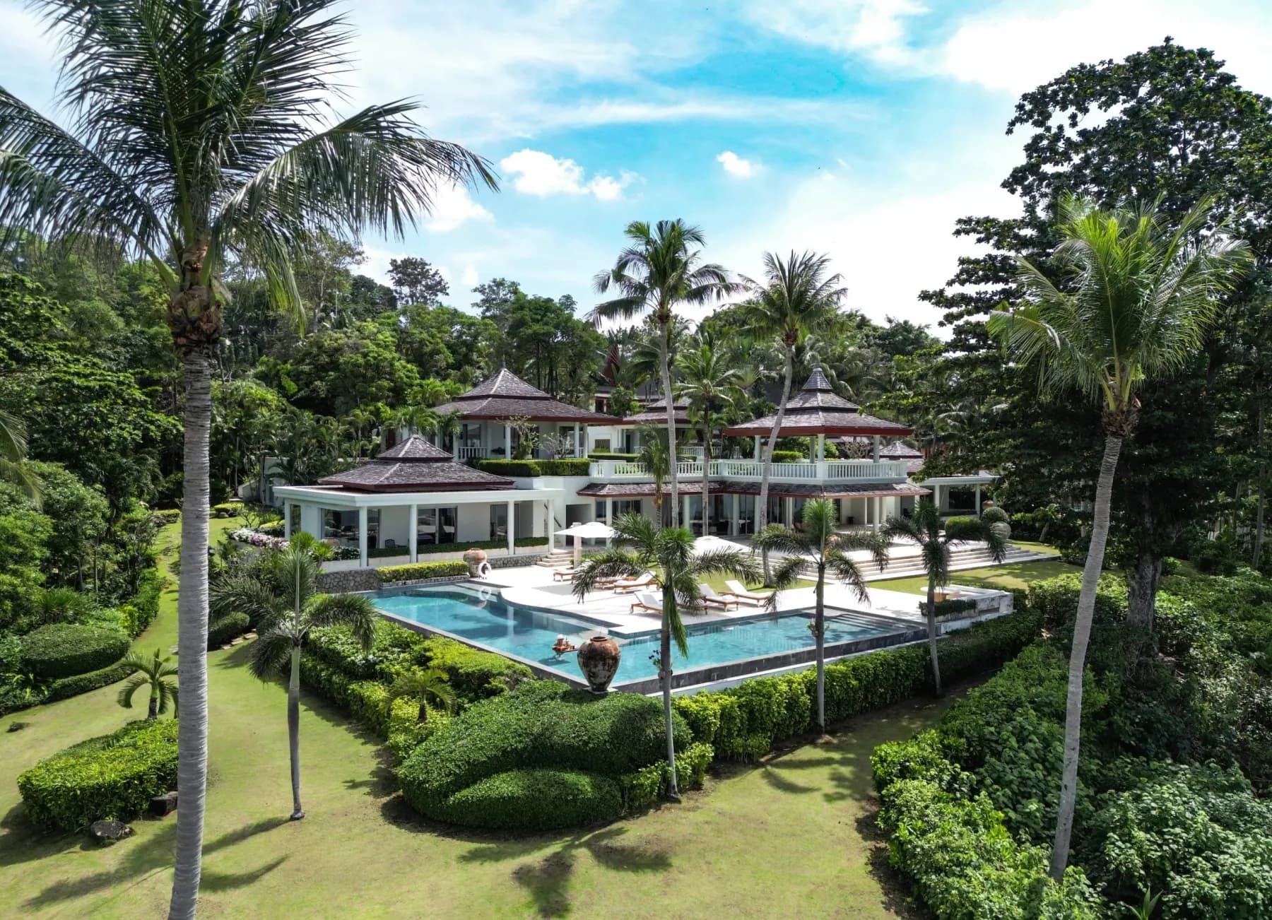 Aerial view of a detached multi-pavilion villa with tiered roofs, L-shaped pool and hedged lawn
