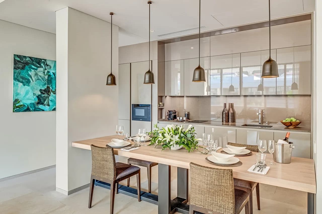 Residence interior: dining table laid for four beside a fitted galley kitchen with bronze pendant lights.