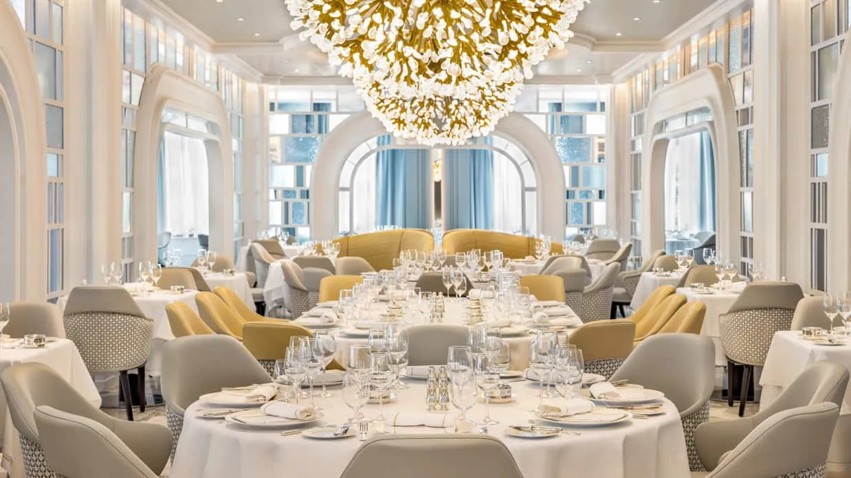 The Grand Dining Room on Oceania Vista, white arches and a large gold chandelier over tables laid for dinner