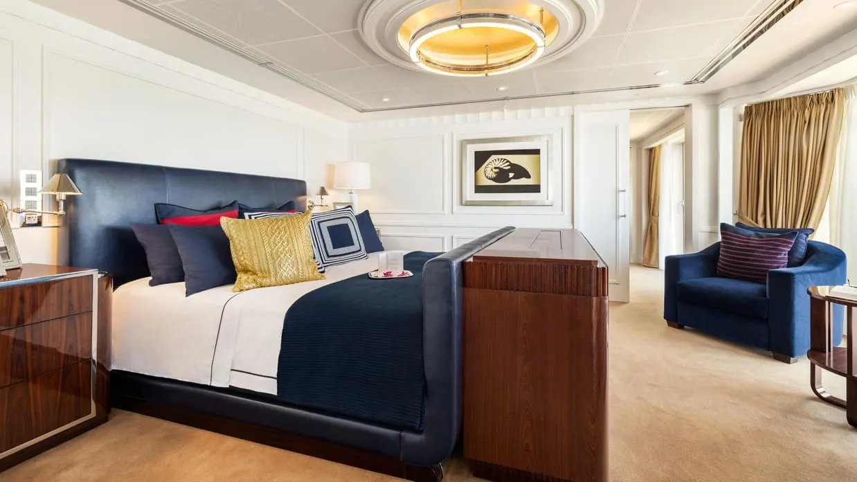The bedroom of the Owner's Suite on Oceania Vista, a navy leather bed, a blue armchair and gold curtains