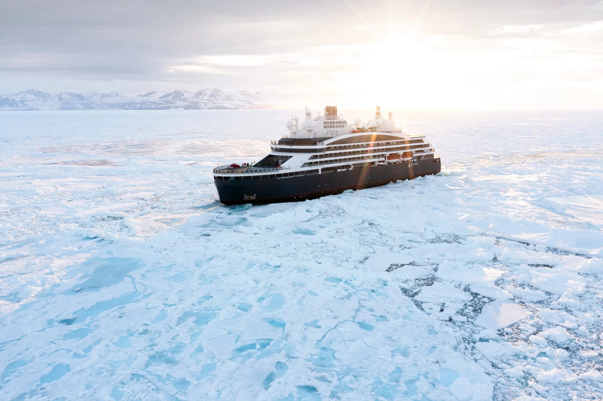 Le Commandant Charcot breaking through polar pack ice towards the camera, snow-covered mountains on the horizon and the sun low behind her