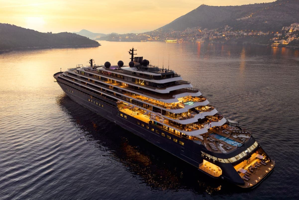 Evrima, the Ritz-Carlton yacht, lit at dusk off a hillside town whose lights are coming on