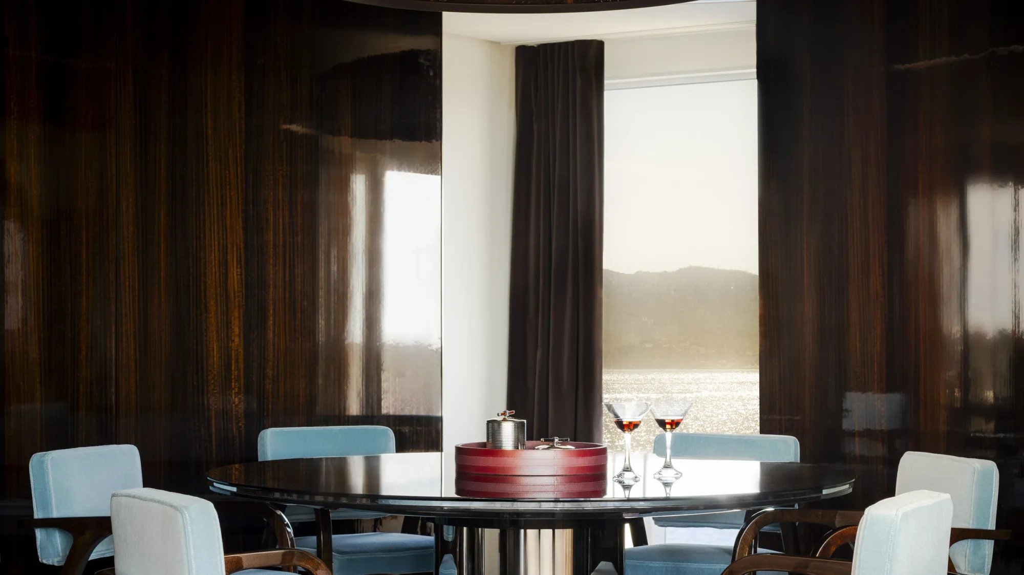 The dining table of the Agatha Christie suite on Orient Express Corinthian, blue chairs and dark lacquered walls beside a window onto the coast