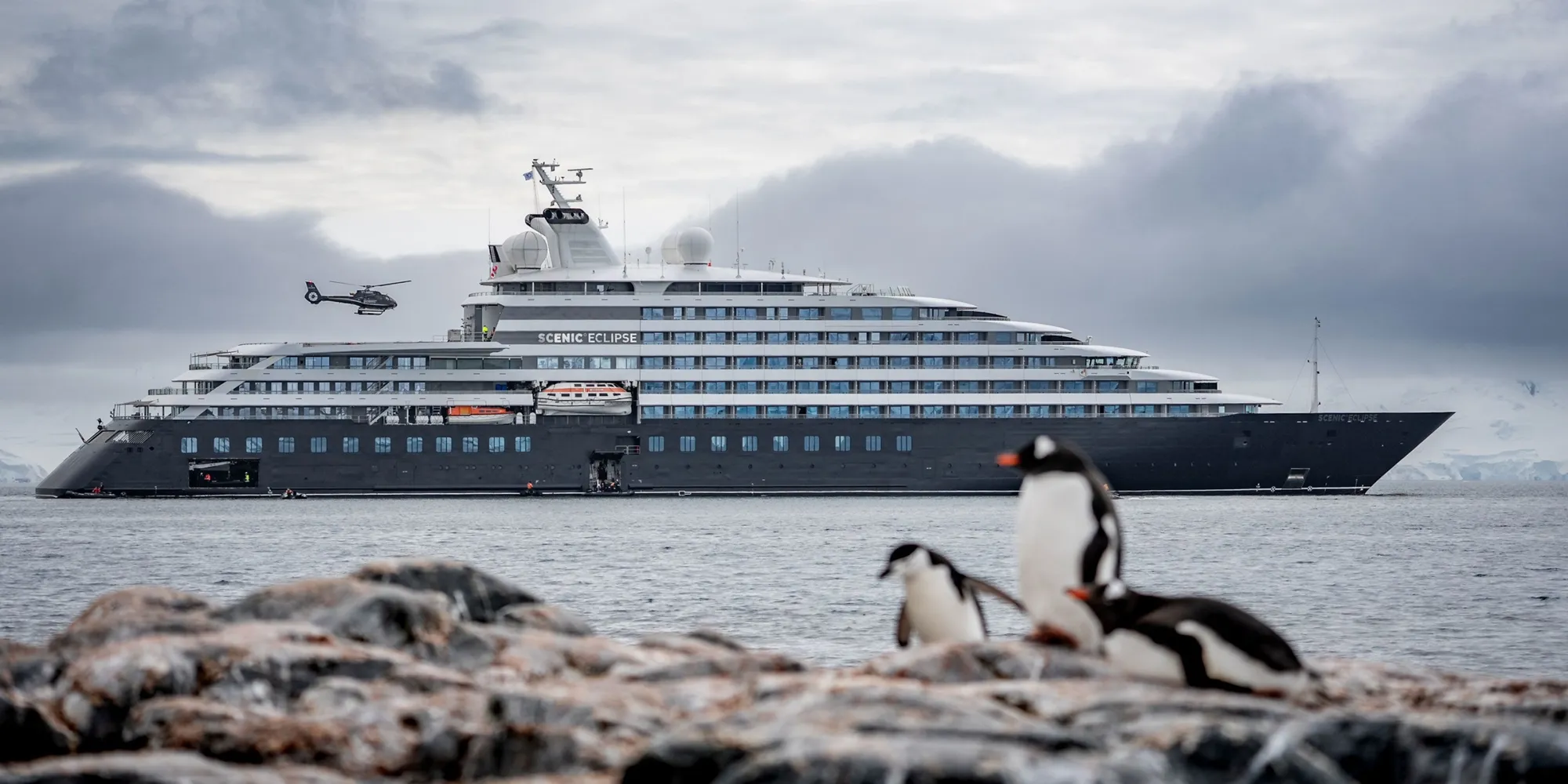 Scenic Eclipse at anchor off Palaver Point, Antarctica, a helicopter in the air beside her and penguins on the rocks in the foreground