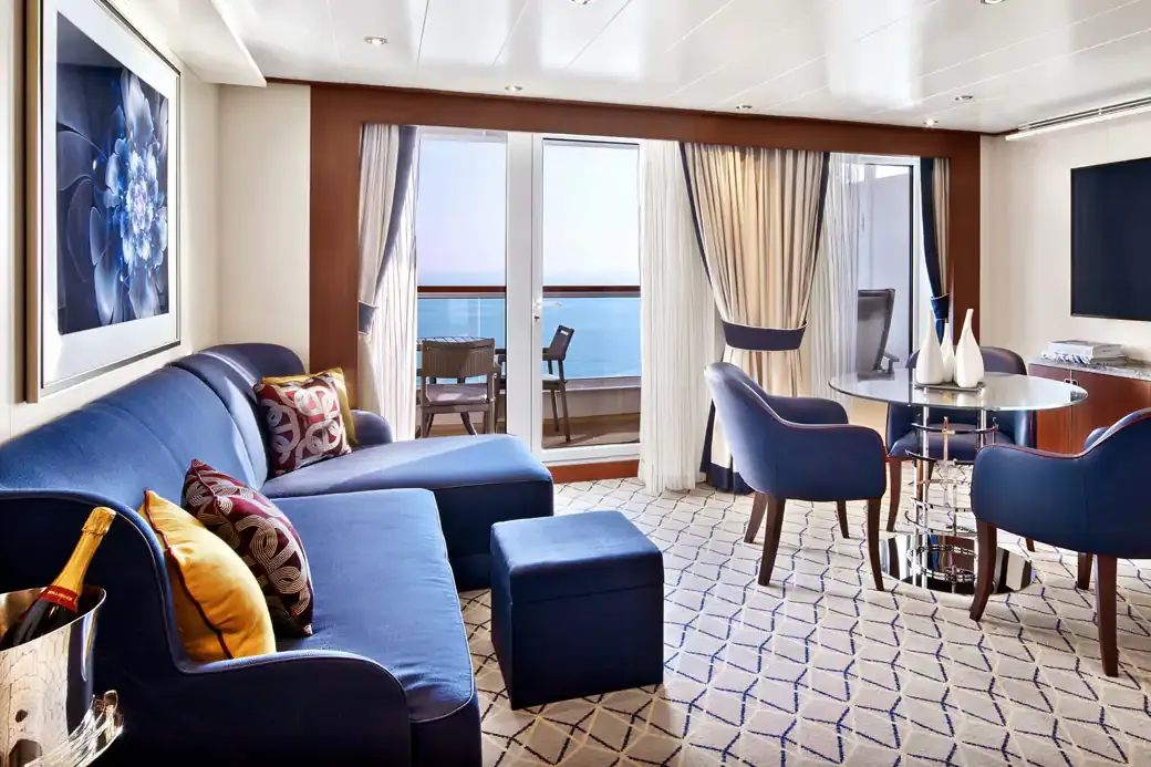 The living room of a Penthouse Suite on Encore and Ovation, a blue sofa and dining chairs before the glass doors to the veranda