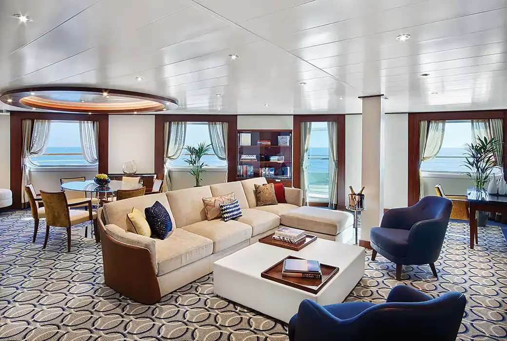 The living room of a Signature Suite on Encore and Ovation, an L-shaped sofa and a dining table under windows on three sides