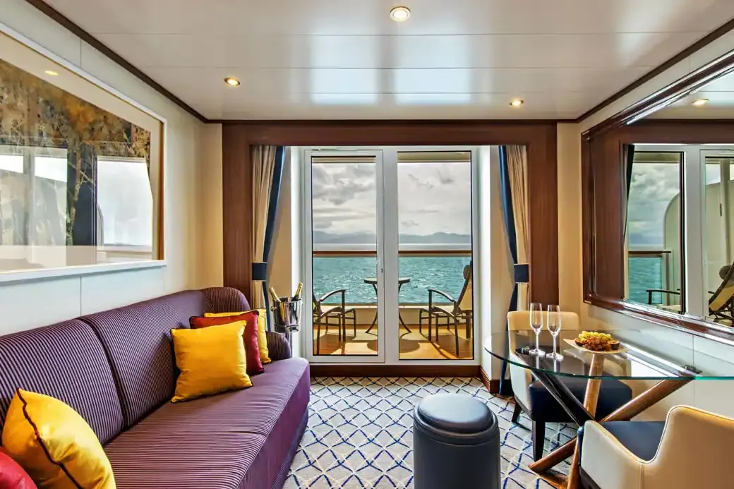 The sitting area of a Veranda Suite on Encore and Ovation, a striped purple sofa and a glass table by the doors to the veranda