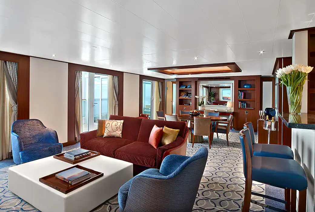 The living room of a Wintergarden Suite on Encore and Ovation, a red sofa, blue armchairs, a bar with stools and a dining table by the windows