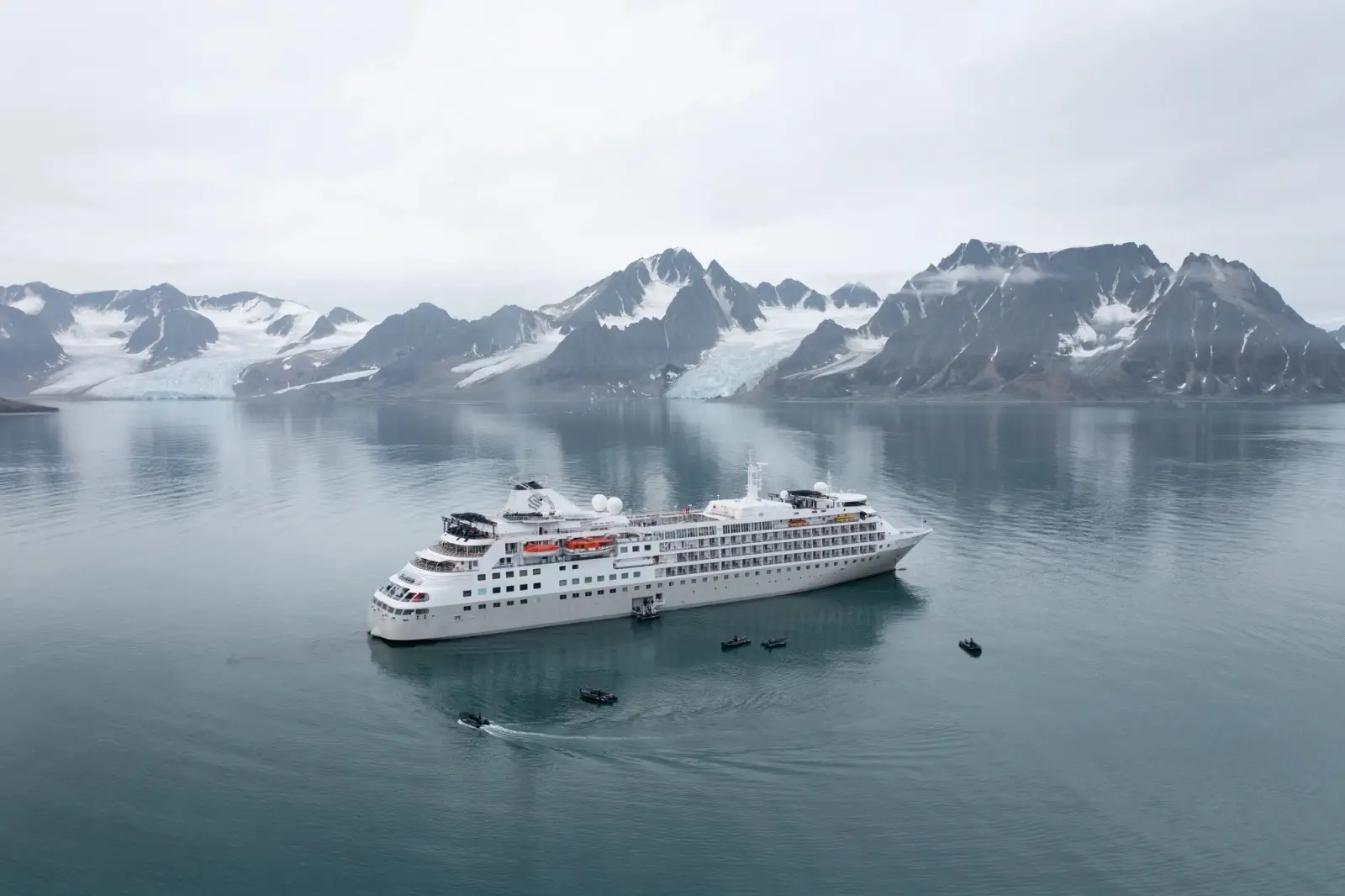 Silver Cloud at anchor in a still bay below glaciers and mountains, Zodiacs around her