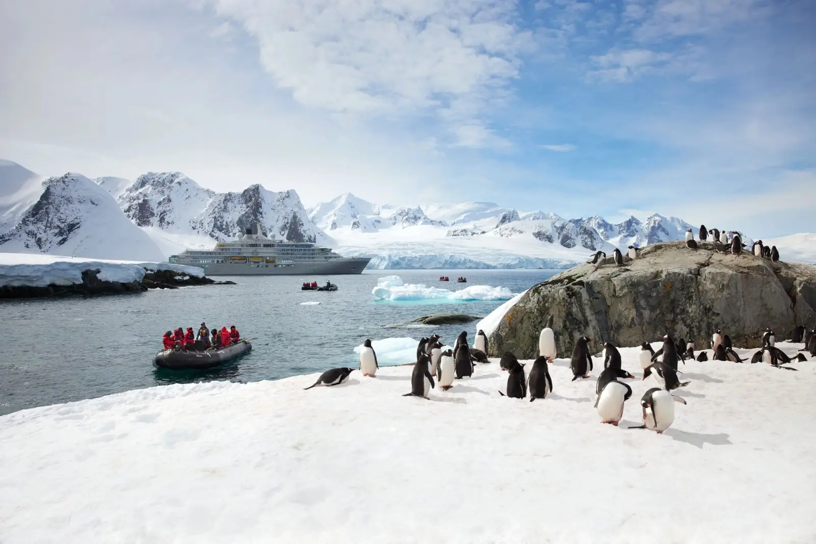 Gentoo penguins on a snowy shore in Antarctica, a Zodiac of guests nearby and Silver Endeavour at anchor beyond