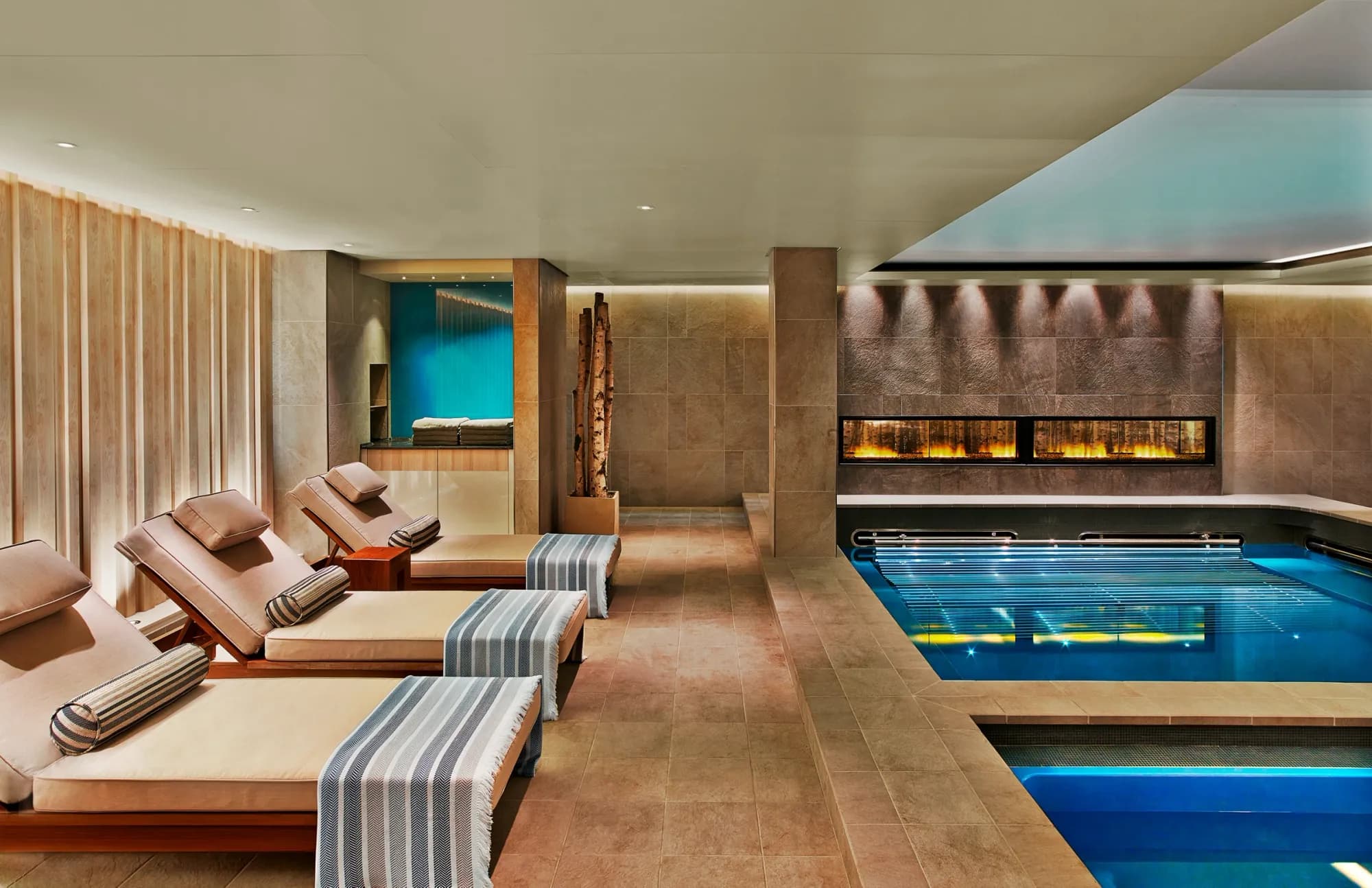 The thermal area of The Nordic Spa, loungers beside a fireplace and a blue-lit pool