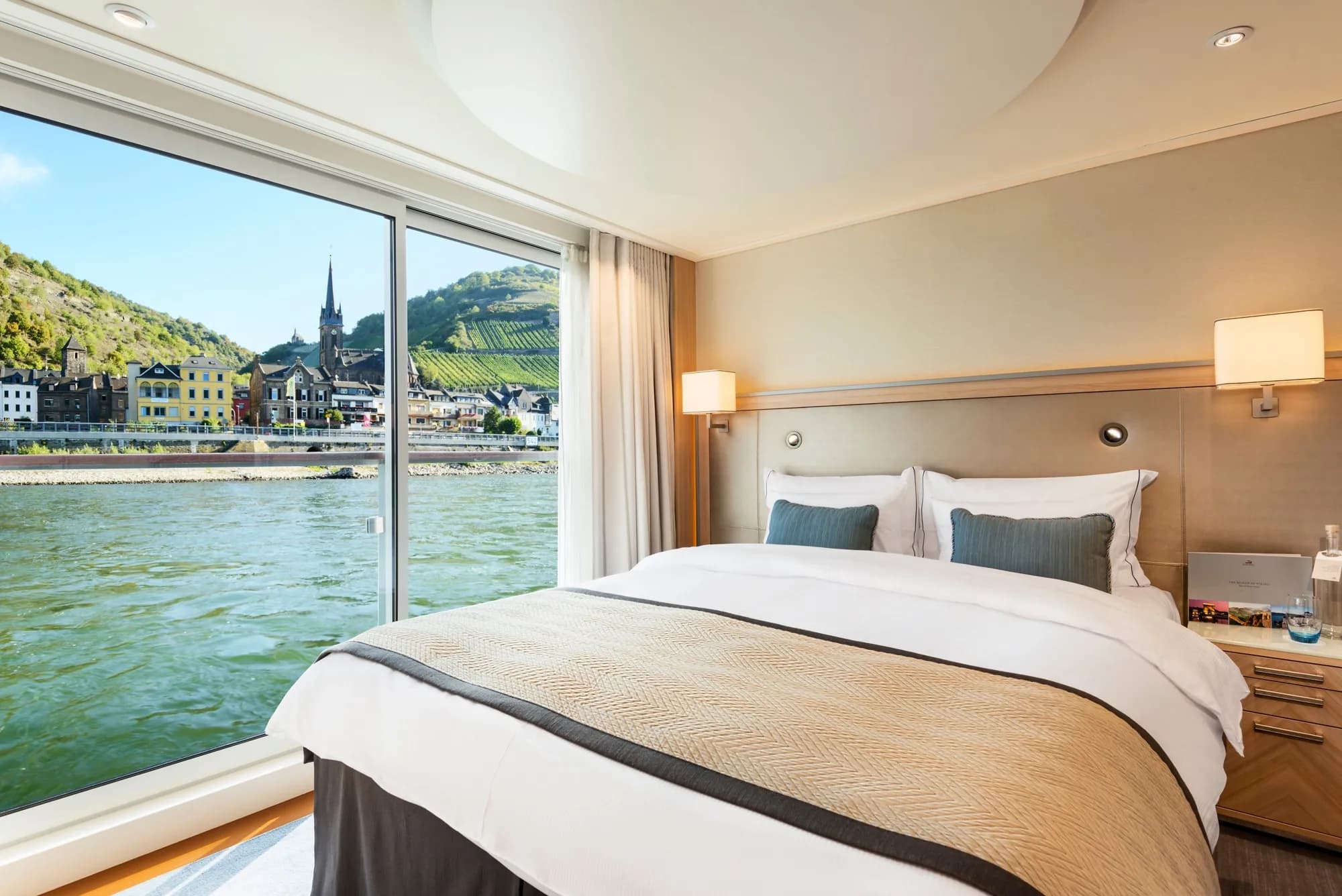 The bedroom of a Veranda Suite on a Viking Longship, a glass door open to the Rhine and a riverside town with vineyards