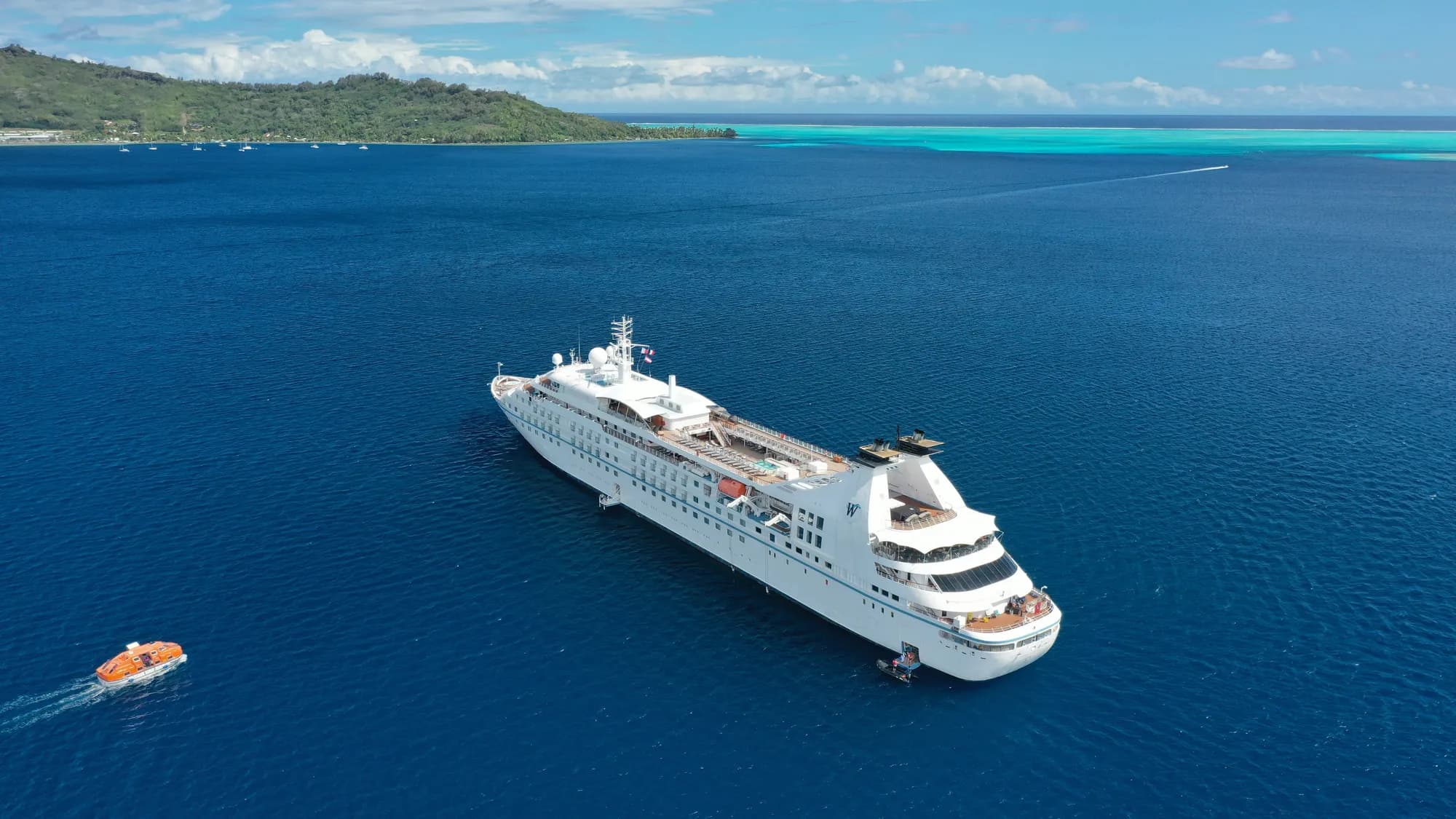 Star Breeze at anchor in deep blue water off a green island with a turquoise lagoon behind