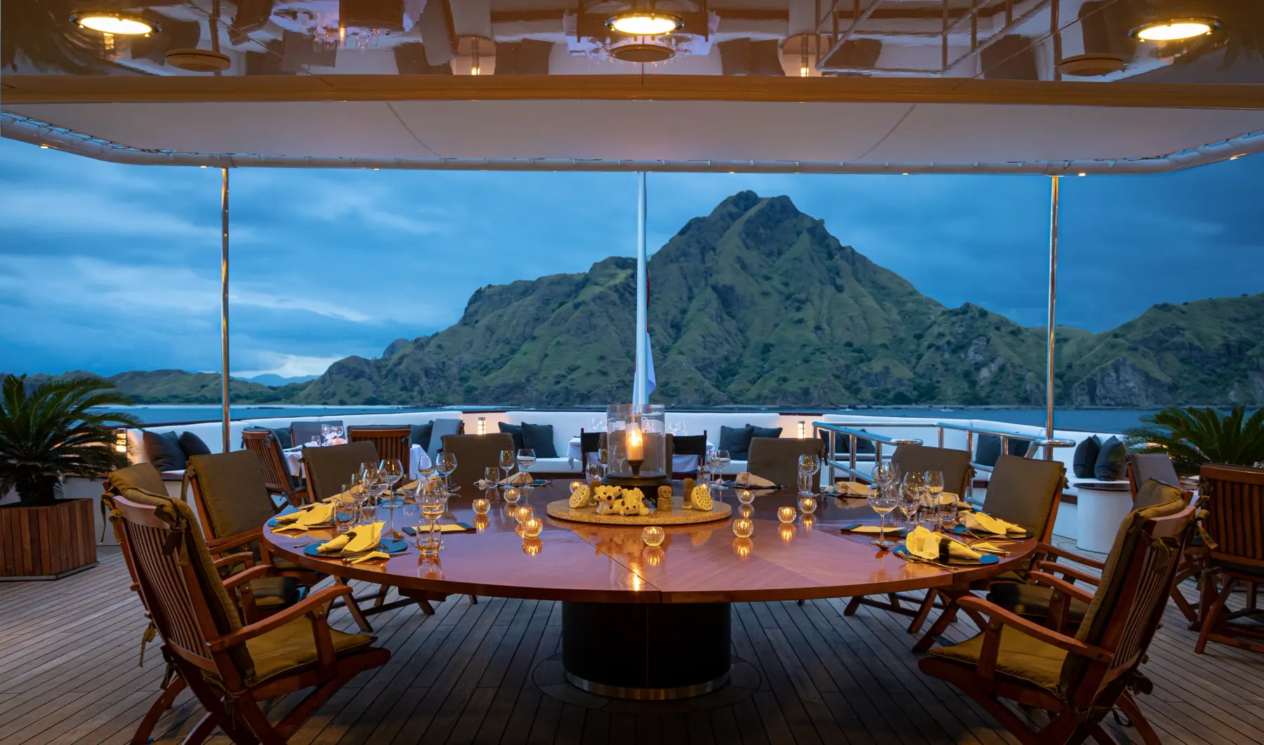 The outdoor dining deck aboard Aqua Blu at dusk, laid for dinner facing a green island peak