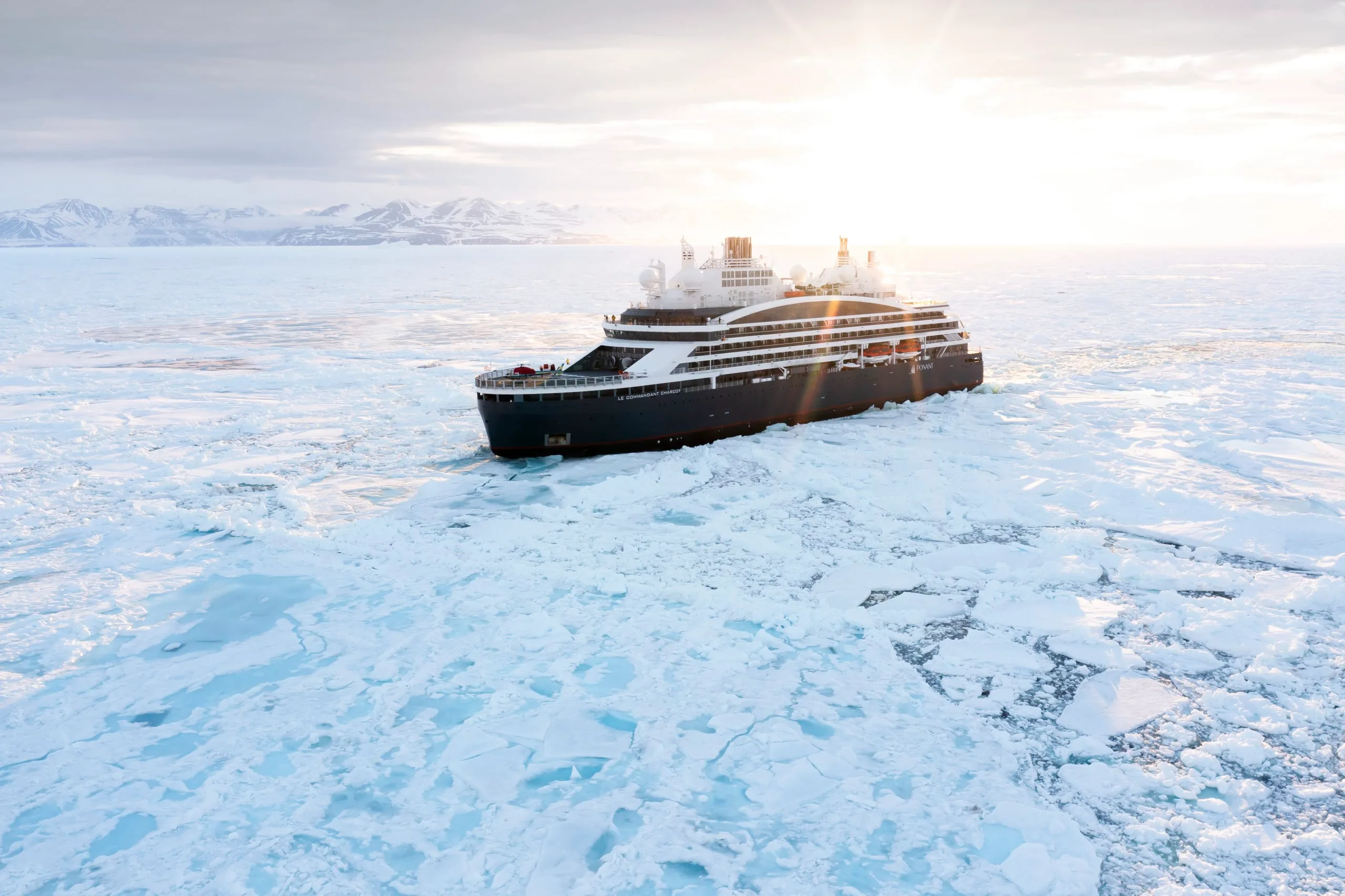 Le Commandant Charcot pushing through polar pack ice under a low sun, mountains on the horizon