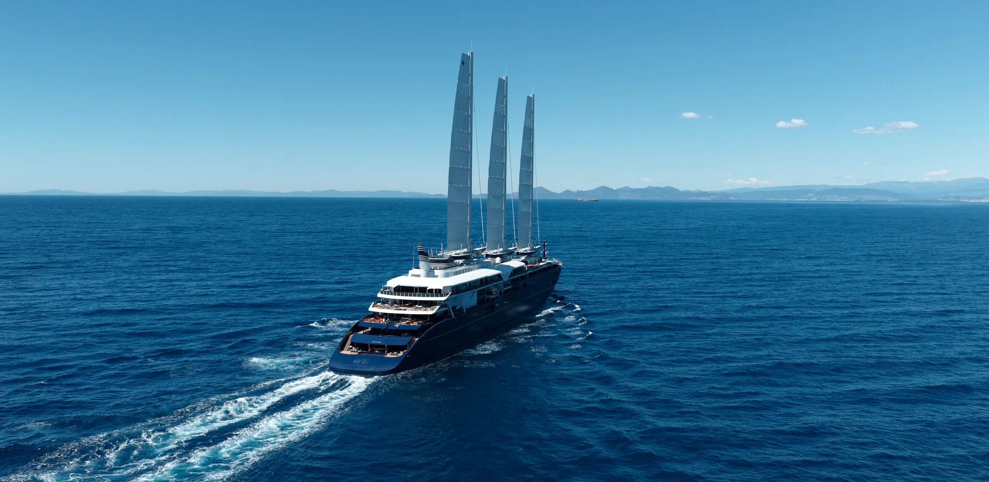 Orient Express Corinthian at sea off a coastline, her three carbon masts raised